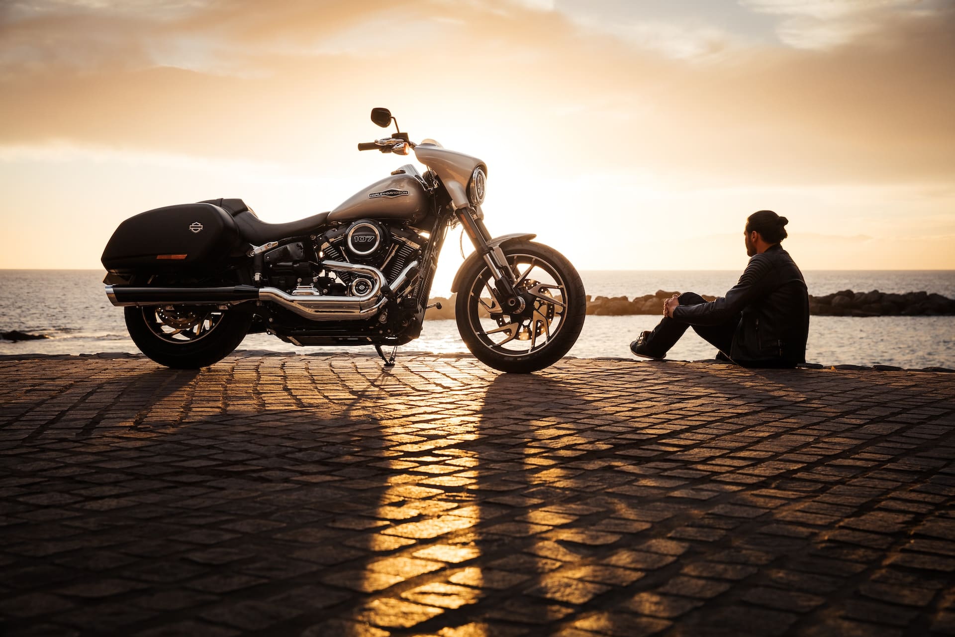 Harley davidson bike next to its rider sitting on a brick road overlooking an ocean view sunset