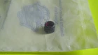 Small dark cylindrical cap with a red center resting on a translucent plastic bag on a bright yellow surface.