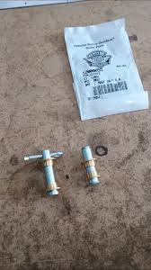 White resealable bag with printed label sits on a brown surface; in front are two small metal valve fittings with blue plastic ends and T-shaped handles, plus a small metal ring between them.