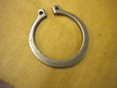 Metal hose clamp (C-shaped) with two tabbed ends on a yellow surface; used to secure hoses or tubing.