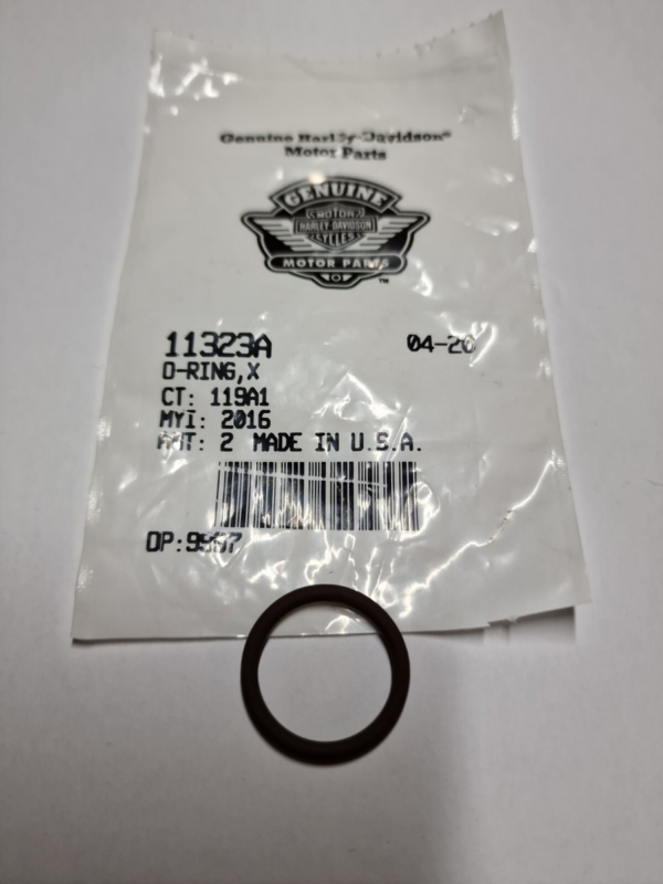Black rubber O-ring resting on a white surface in front of a plastic Harley-Davidson Genuine Motor Parts bag showing part number 11323A and barcode label