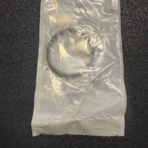 Sealed clear plastic bag containing a single black circular rubber O-ring on a dark textured surface