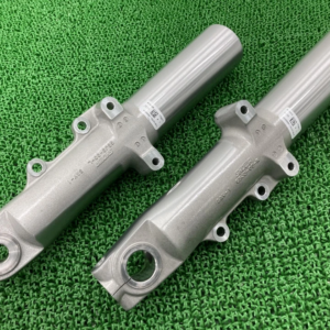 Two gray motorcycle front fork legs with mounting tabs on a green textured surface, shown side by side.
