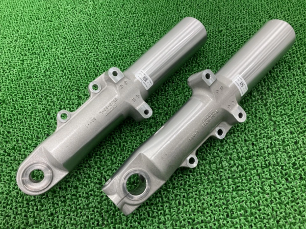 Two gray motorcycle front fork legs with mounting tabs on a green textured surface, shown side by side.