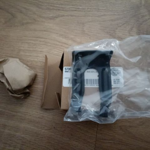 Black plastic mounting bracket in a clear plastic bag, placed on a wooden floor beside brown packing paper and a small cardboard box.