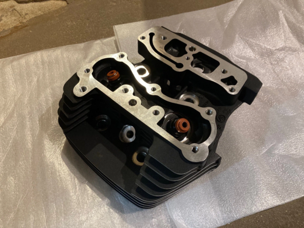 Motorcycle or small engine cylinder head with aluminum gasket, cooling fins, and orange valve seals placed on a protective sheet.