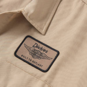 Close-up of a beige work shirt featuring a Dickies patch with wings and the words 'Built to Outlast' on tan fabric