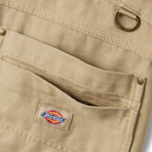 Close-up of a beige Dickies work backpack pocket with a zipper and metal D-ring detail on canvas fabric.