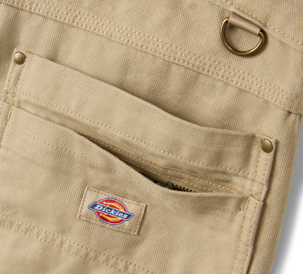 Close-up of a beige Dickies work backpack pocket with a zipper and metal D-ring detail on canvas fabric.