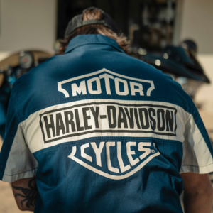 Person wearing a blue Harley‑Davidson jacket with logos on the back, viewed from behind at a motorcycle showroom or outdoor area.