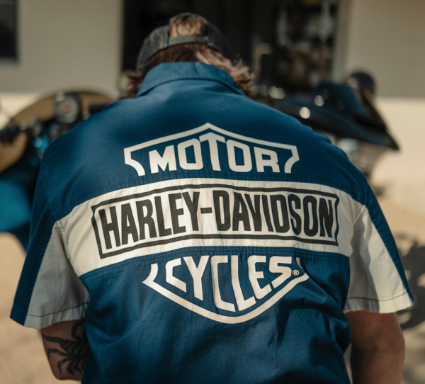 Person wearing a blue Harley‑Davidson jacket with logos on the back, viewed from behind at a motorcycle showroom or outdoor area.