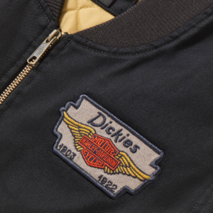 Close-up of a black jacket with a Dickies patch featuring wings and a Harley‑Davidson emblem.