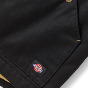 Close-up of a black Dickies work jacket with a brass button, showing the small brand label near the hem.