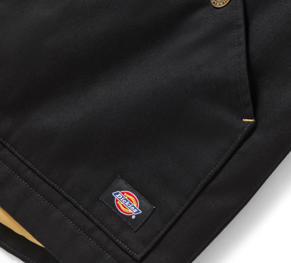 Close-up of a black Dickies work jacket with a brass button, showing the small brand label near the hem.